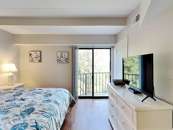 A bedroom with a bed, a TV, a dresser, and a balcony with a view of trees.