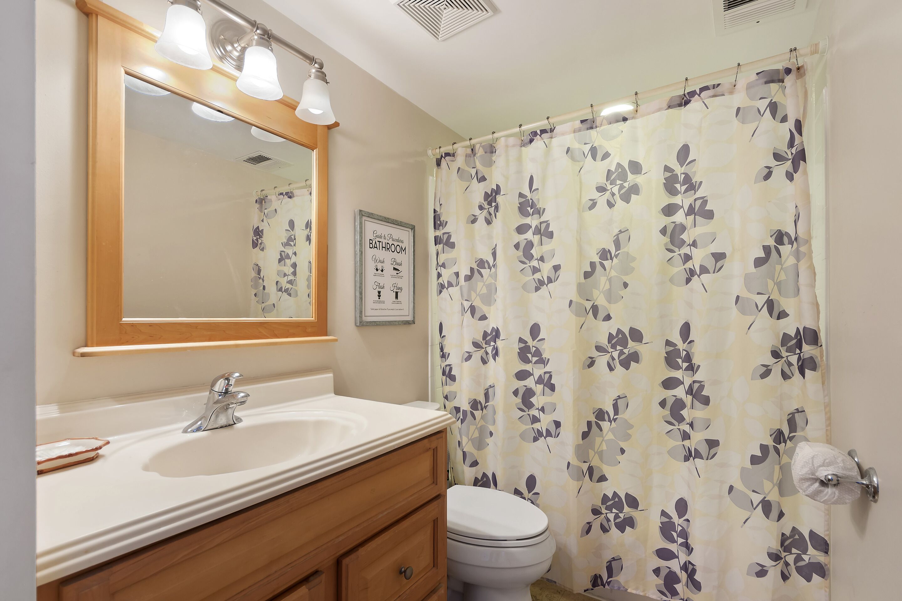 A bathroom with a white sink, wooden vanity, a mirror with lights, a toilet, and a floral shower curtain.