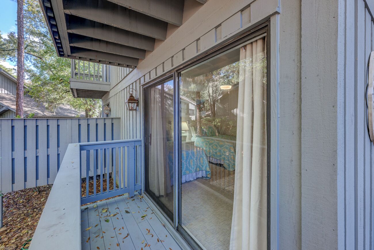 A covered patio with a blue railing, a sliding glass door leading to a room with a bed and a table, and a wooden deck with scattered leaves.