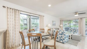 A spacious living area with a dining table, chairs, a sofa, and a balcony with curtains. - Hickory Cove 47 by Hilton Head Retreats (Hilton Head Island)