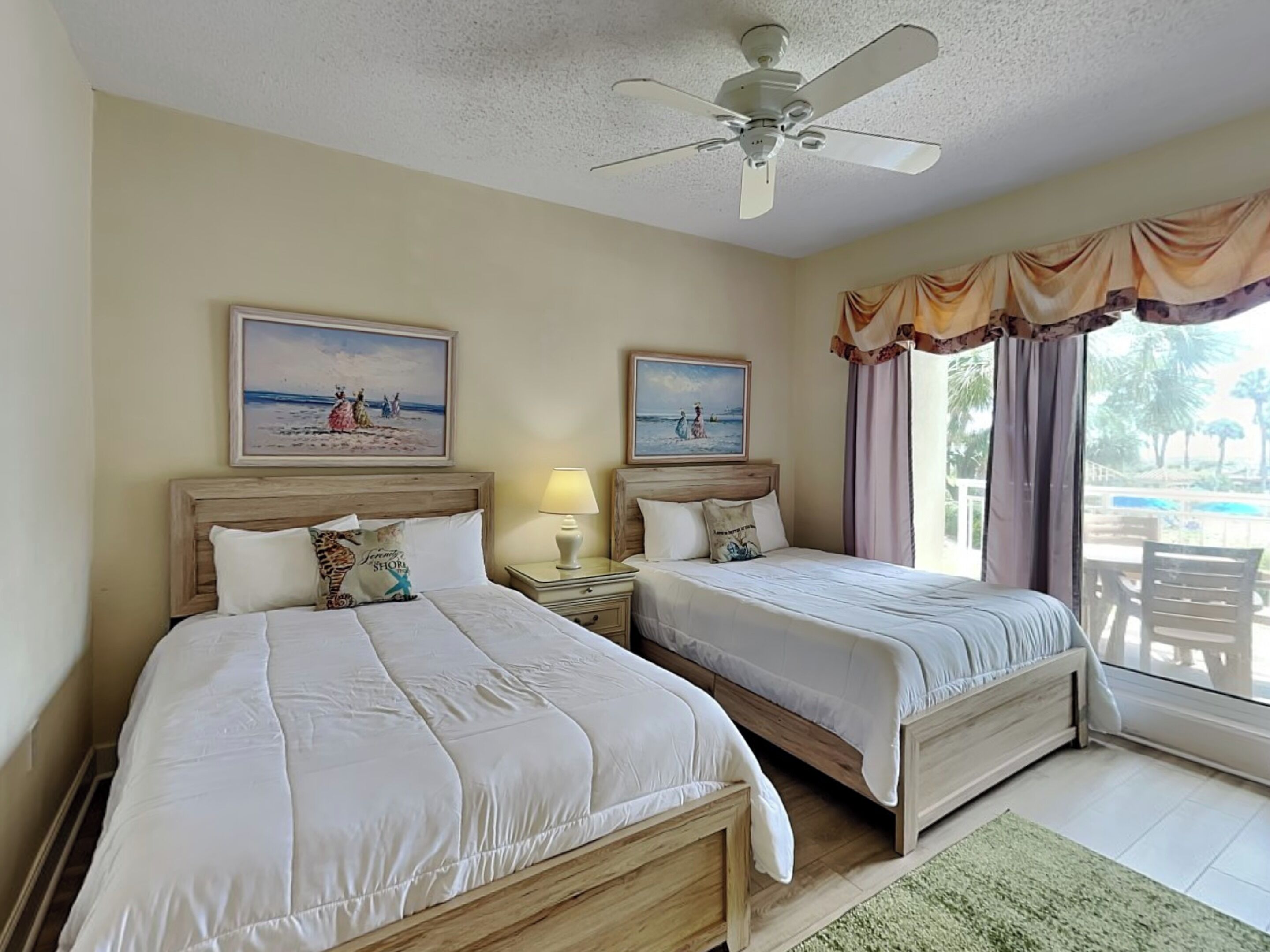A hotel room with two beds, a ceiling fan, a window with curtains, and artwork on the walls.