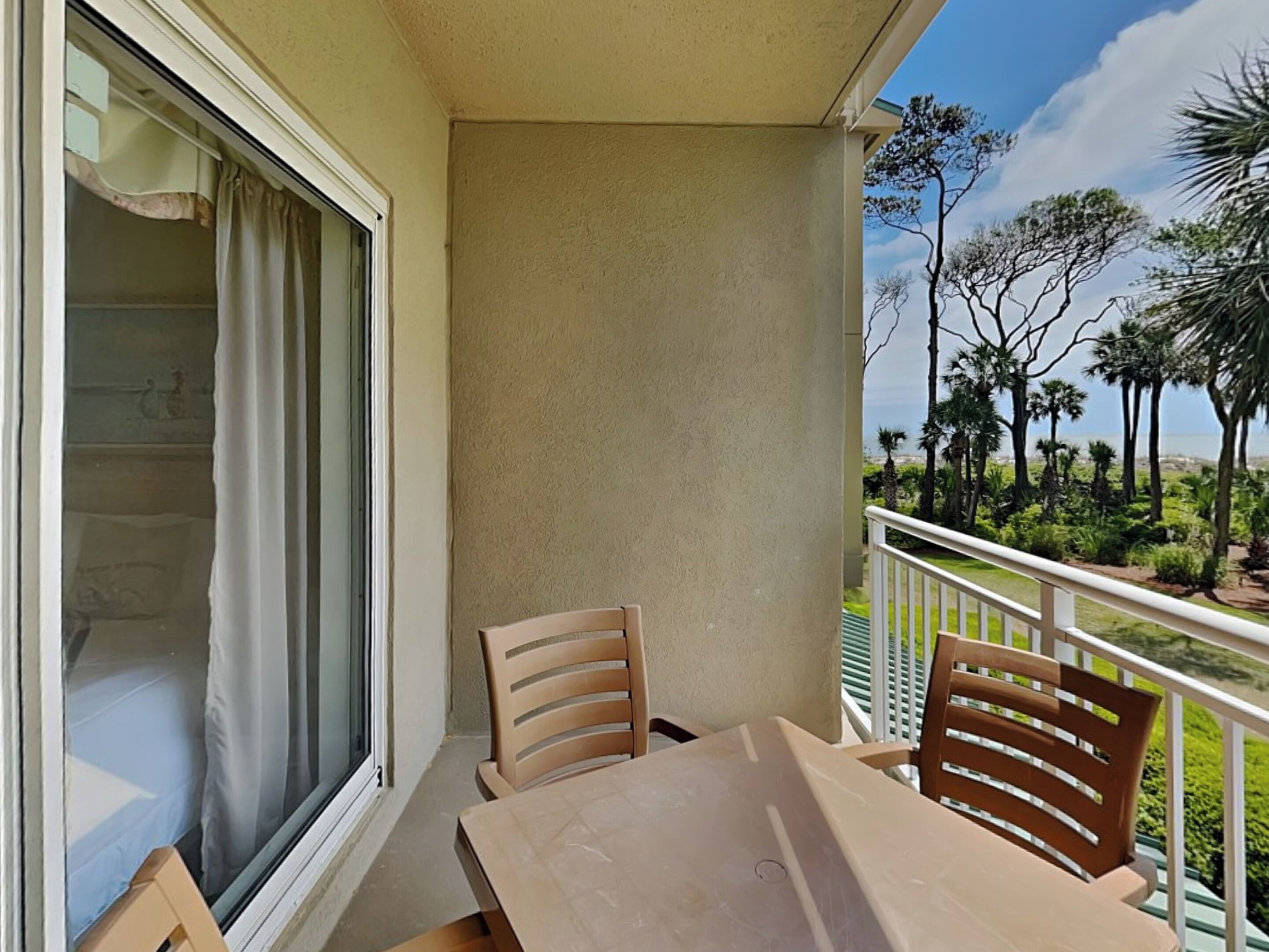 A balcony with a table and chairs, overlooking a landscaped area with trees and a view of the ocean.