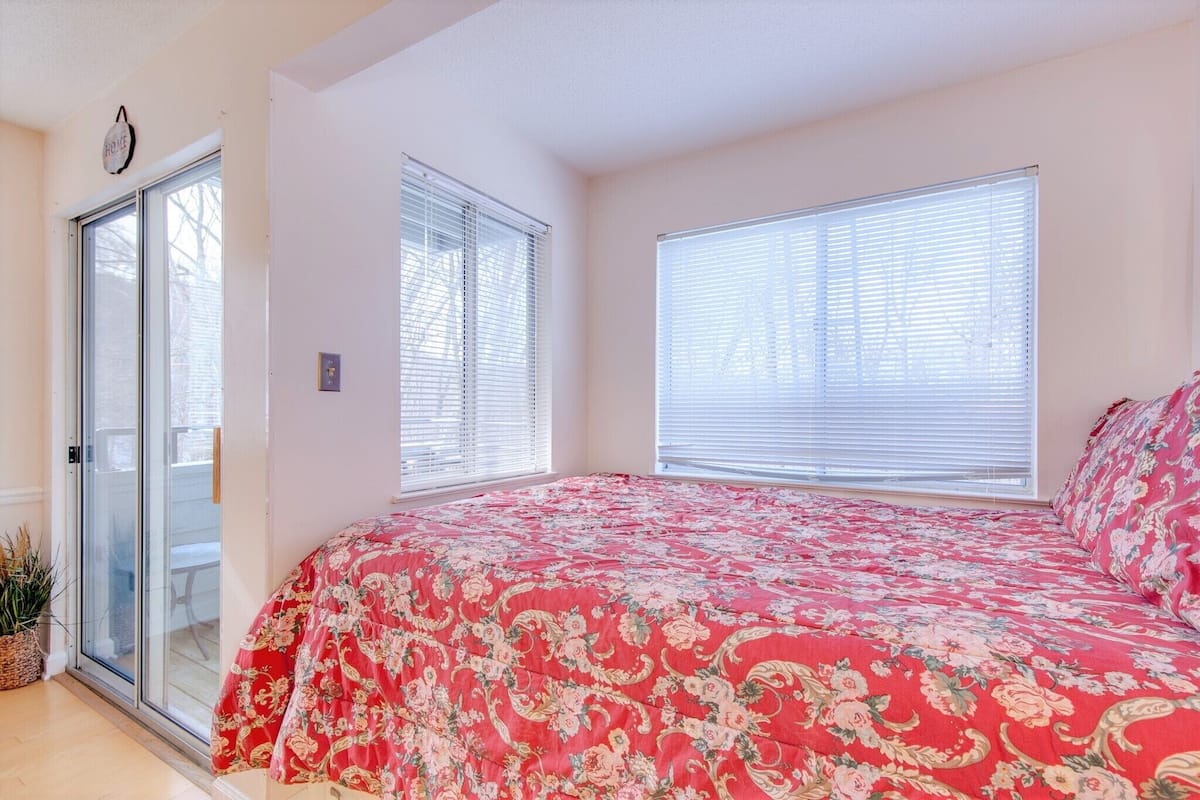 A bedroom with a large bed, two windows with blinds, a sliding glass door, and a small plant.