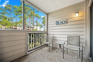 A balcony with two chairs and a table, a 'BEACH' sign, and a view of trees and buildings.