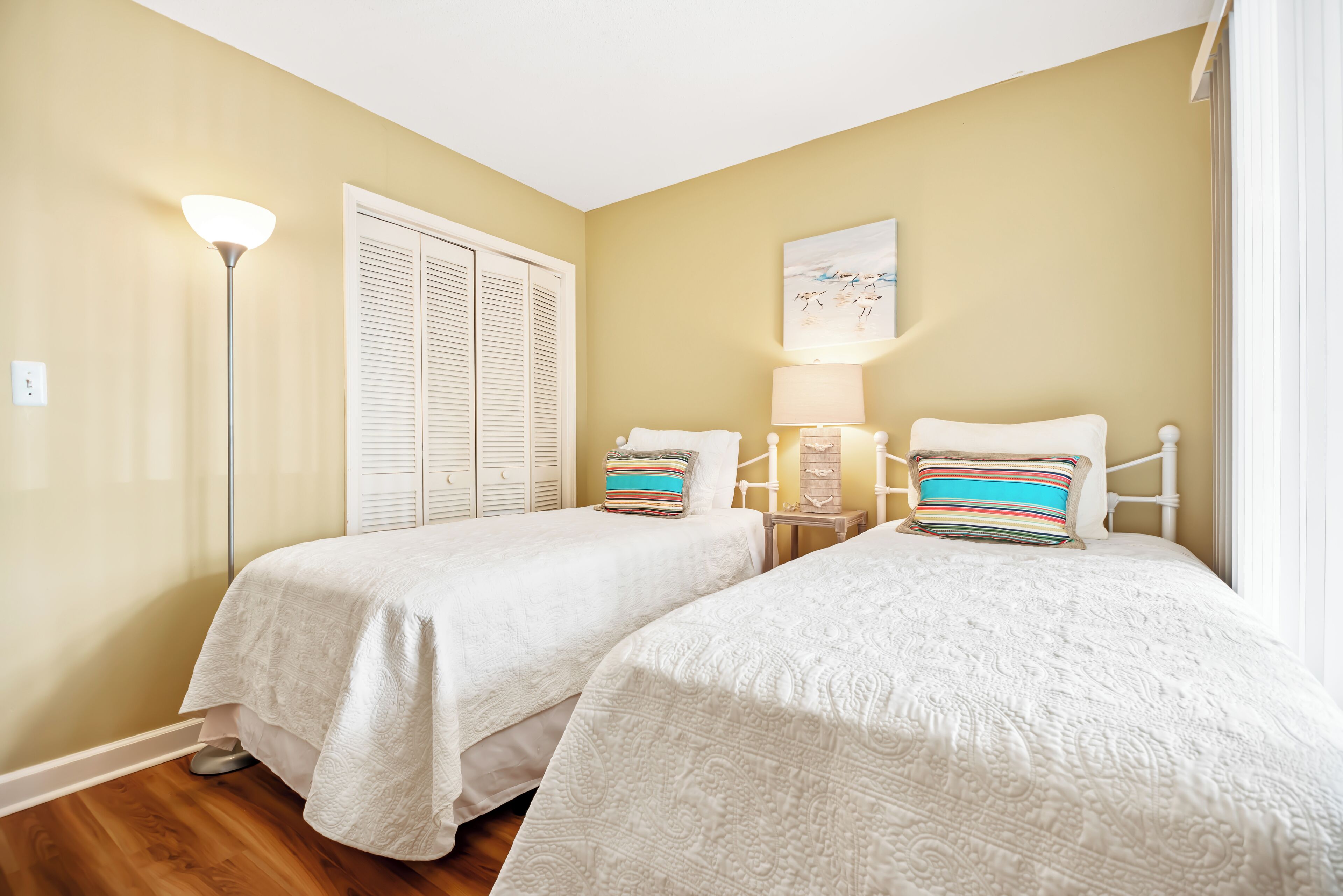 A bedroom with two beds, a bedside lamp, a window with blinds, and a wall-mounted artwork.