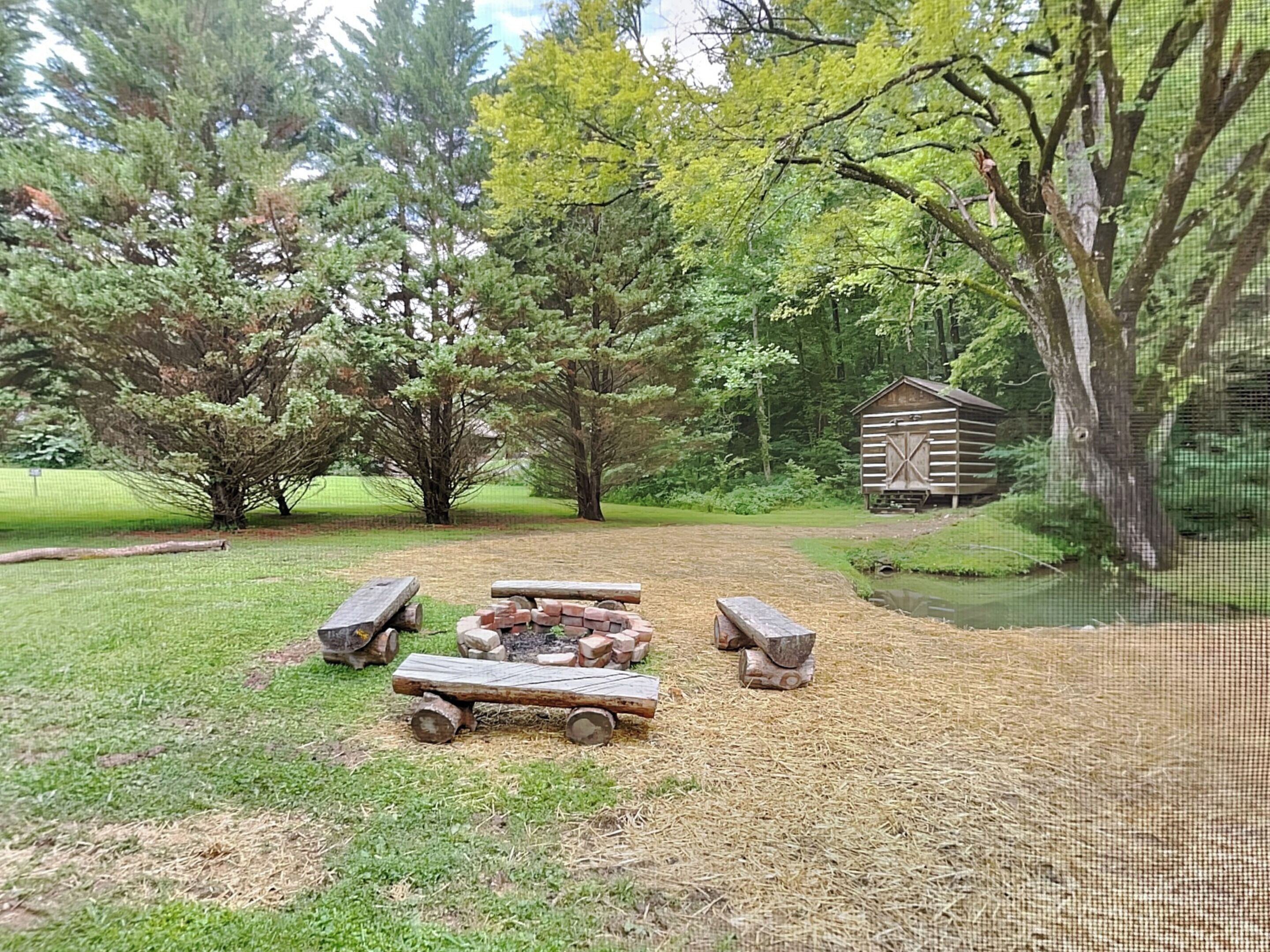Un espacio de estar al aire libre rústico con una chimenea, rodeado de árboles y una estructura de madera al fondo.
