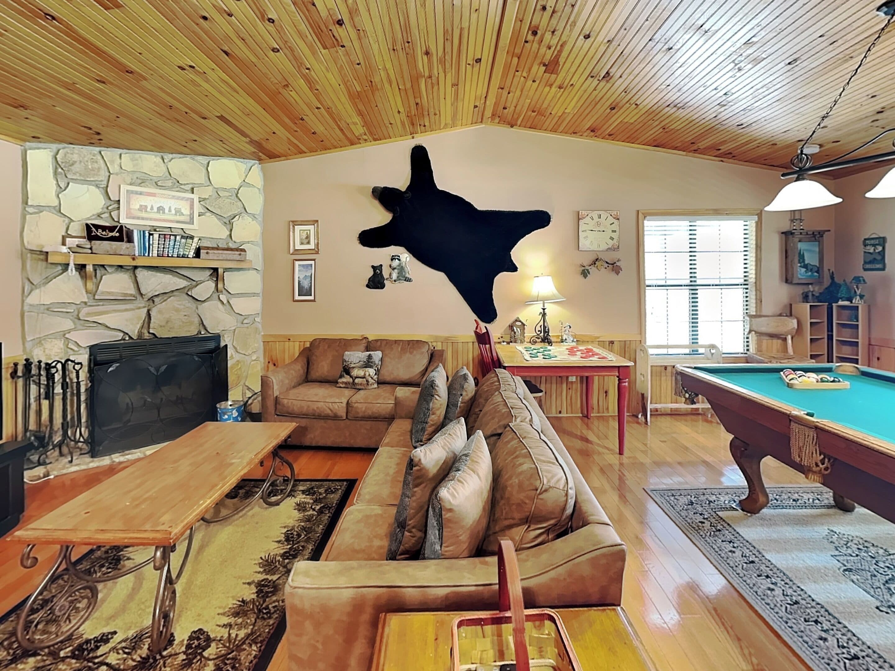 A cozy living room with a stone fireplace, wooden ceiling, a pool table, and a large bear sculpture.