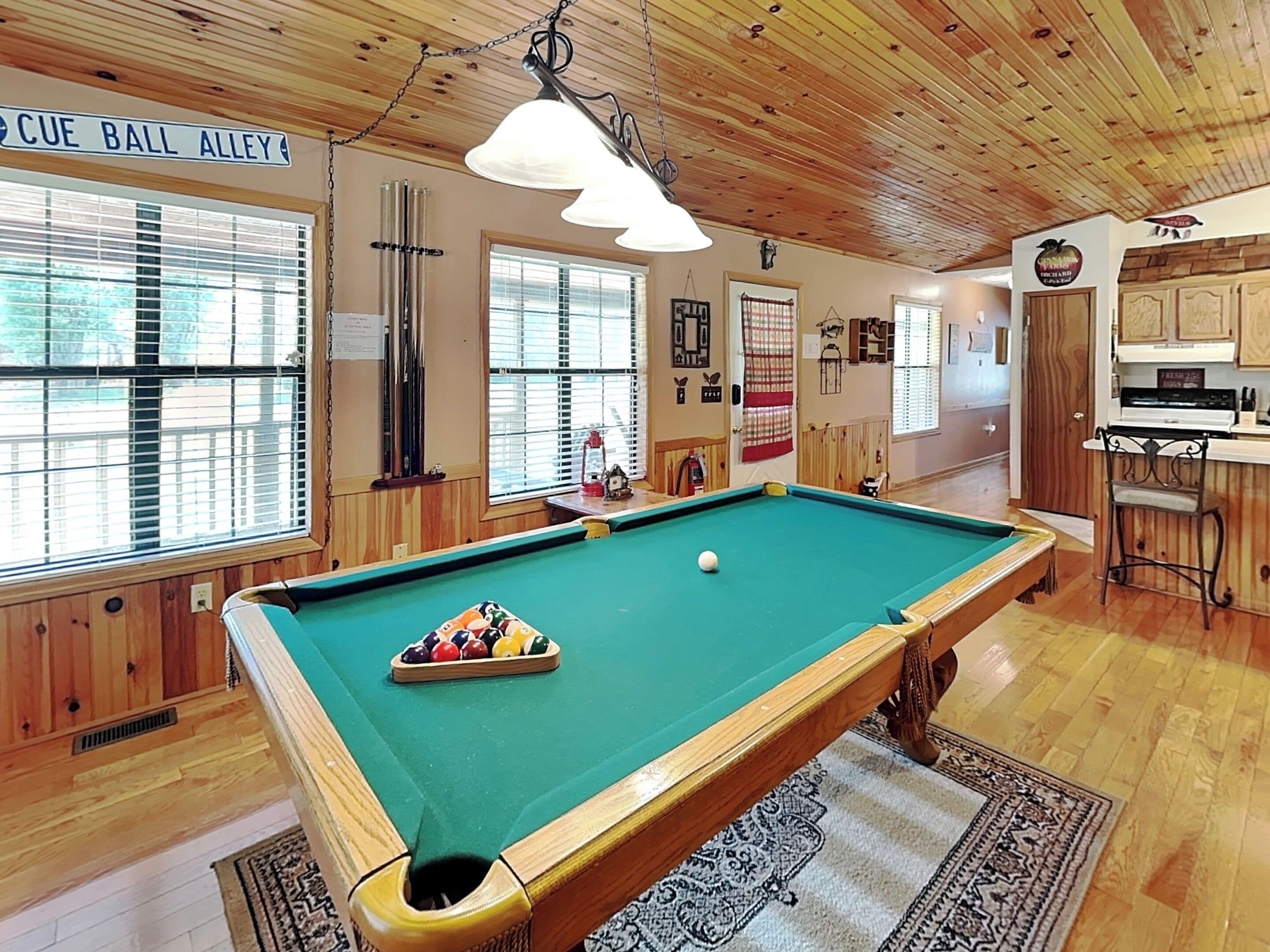 Una mesa de billar en una habitación con paredes y techo de madera, una zona de bar y una ventana con persianas.