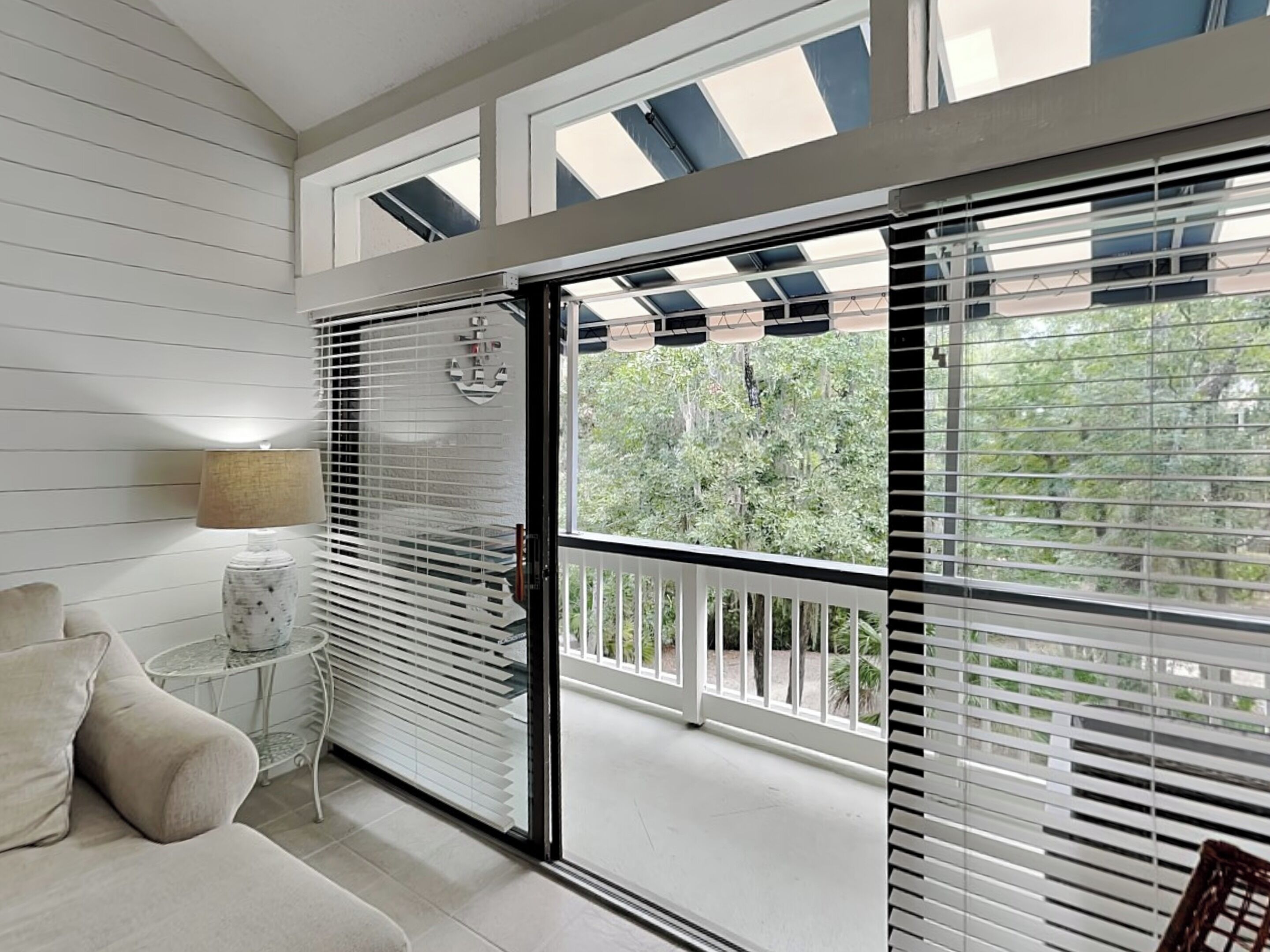 A room with a sliding glass door leading to a balcony, a small round table with a lamp, and a sofa.