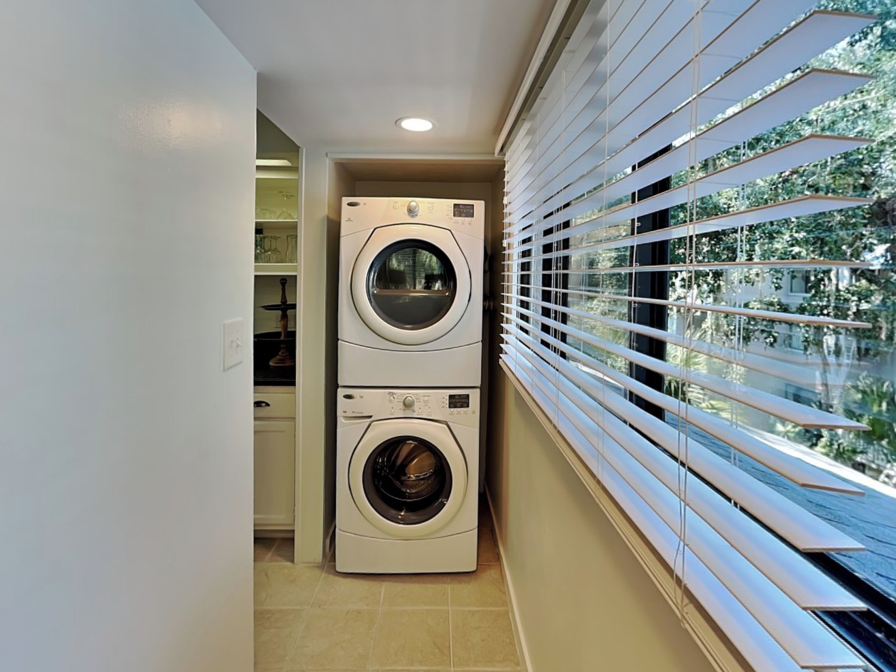 A laundry room with two stacked washing machines, a window with blinds, and a tiled floor.