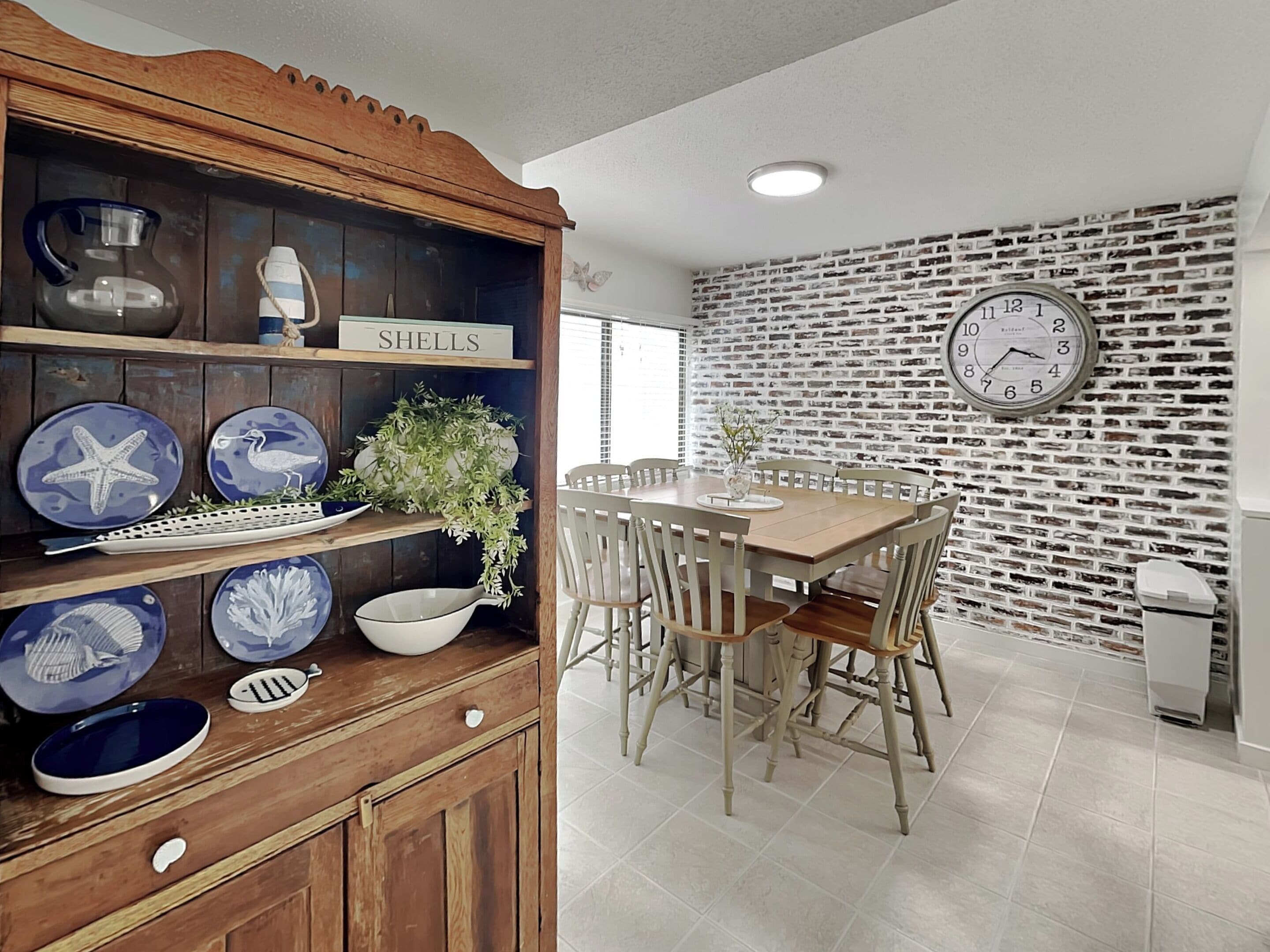 A dining area with a wooden cabinet displaying decorative plates, a brick-patterned wall, and a clock.