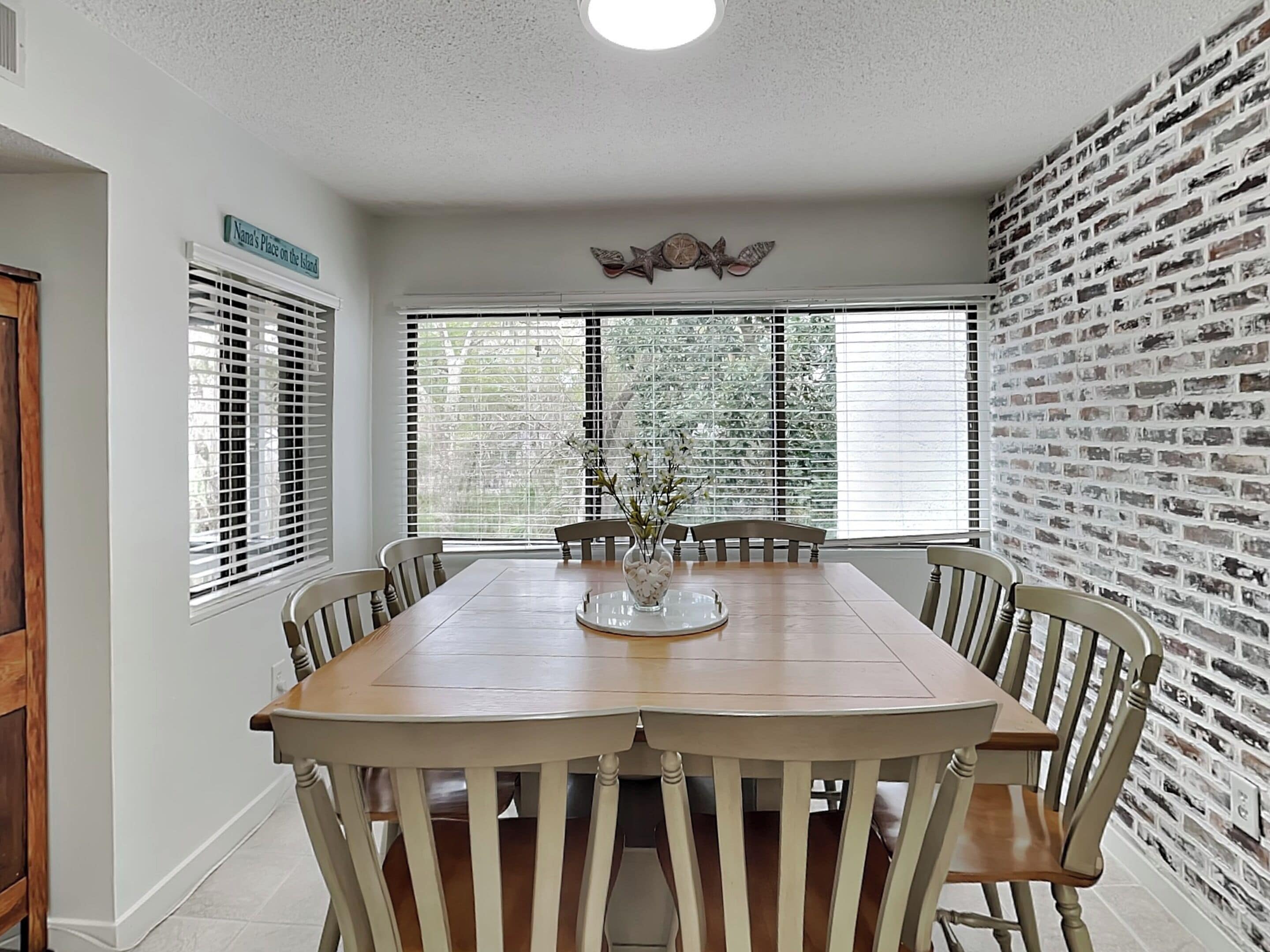 Un comedor con una mesa y sillas de madera, ventanas grandes con persianas, una pared de ladrillo y un letrero en la pared.