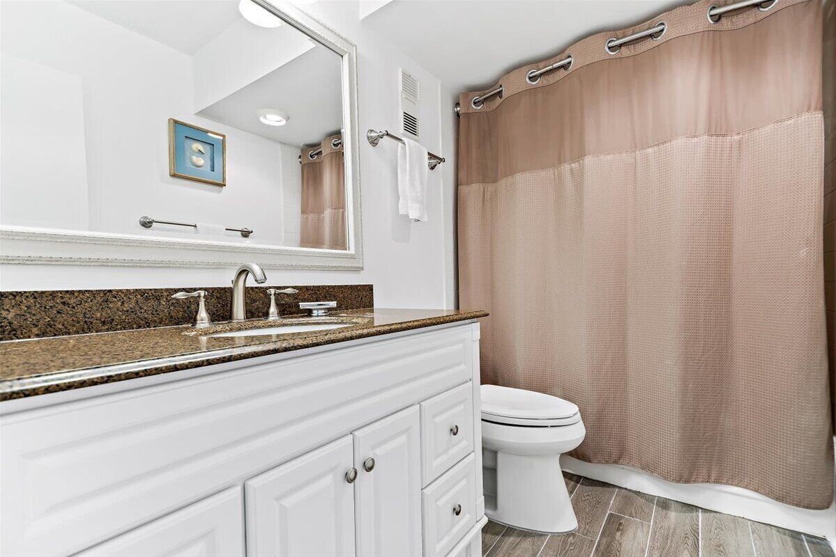 A bathroom with a white vanity, a large mirror, a sink with a granite countertop, a toilet, and a beige shower curtain.