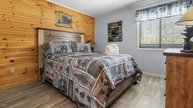 A bedroom with a wooden headboard, a patterned bedspread, a nightstand, and a window with blinds. - Bear Cub Retreat by Jackson Mountain Rentals (Gatlinburg)