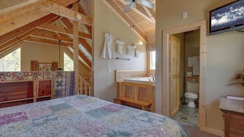 A bedroom with a large bed, wooden ceiling beams, a ceiling fan, a wooden bench, a quilt, a bathroom with a toilet and sink, and a TV mounted on the wall.