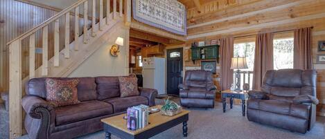 A cozy log cabin interior with a brown leather sofa, two leather armchairs, a wooden coffee table, and a staircase leading to an upper level.