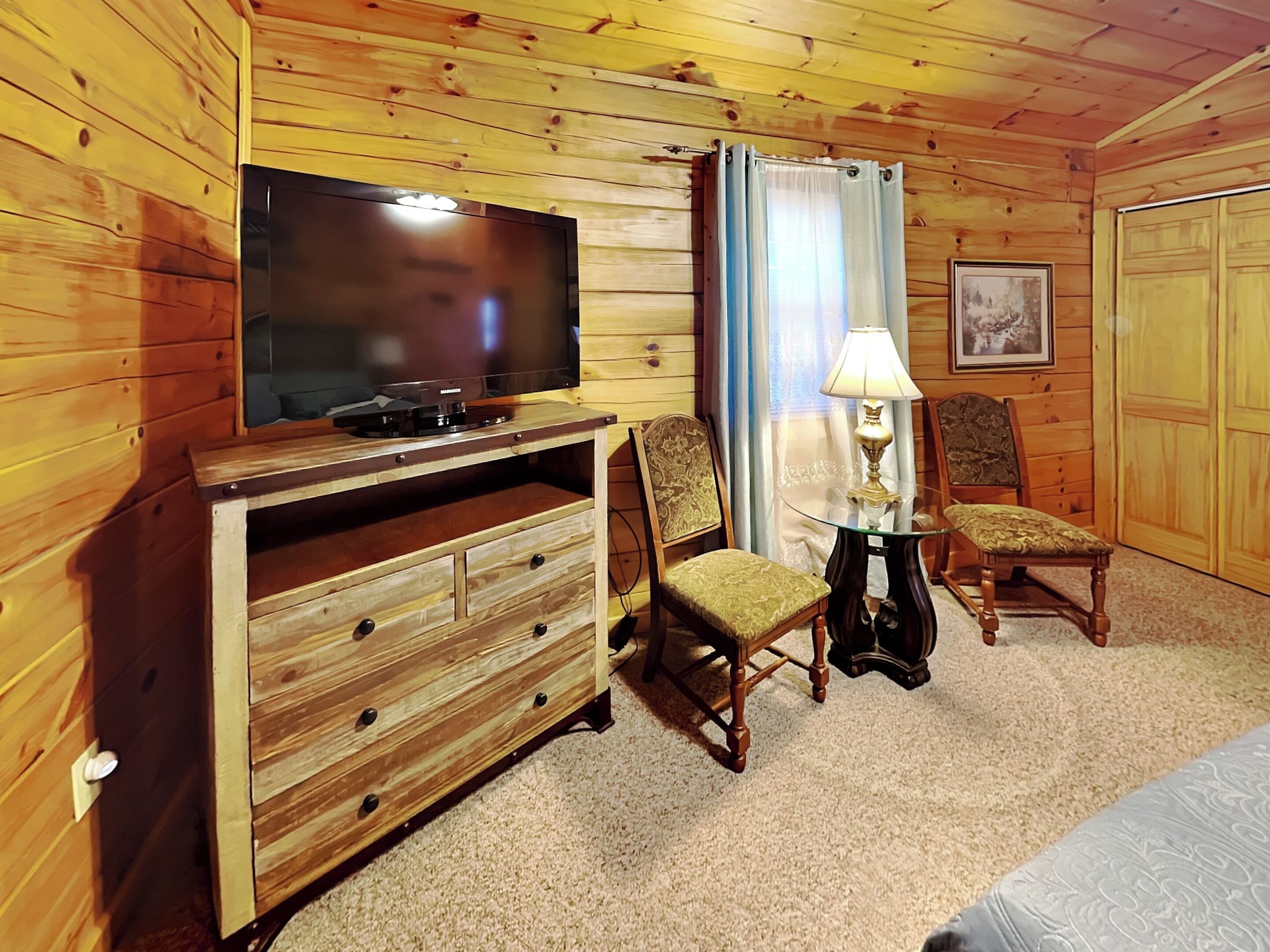 Una habitación con paneles de madera, un televisor sobre un mueble de madera, dos sillones tapizados, una mesita con una lámpara y un cuadro enmarcado en la pared.