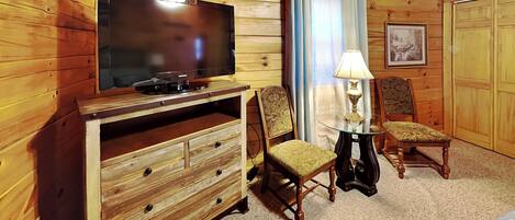 Una habitación con paneles de madera, un televisor sobre un mueble de madera, dos sillones tapizados, una mesita con una lámpara y un cuadro enmarcado en la pared.