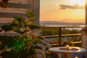 Un balcón con vistas a la puesta de sol sobre un cuerpo de agua, una planta en maceta en primer plano y una pequeña mesa con una fuente.