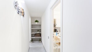 A narrow hallway with white walls and tiled flooring leading to a kitchen area with a dining table and chairs. Shelves with plants and decorative items are visible. - Nùe Rooms (Cagliari)
