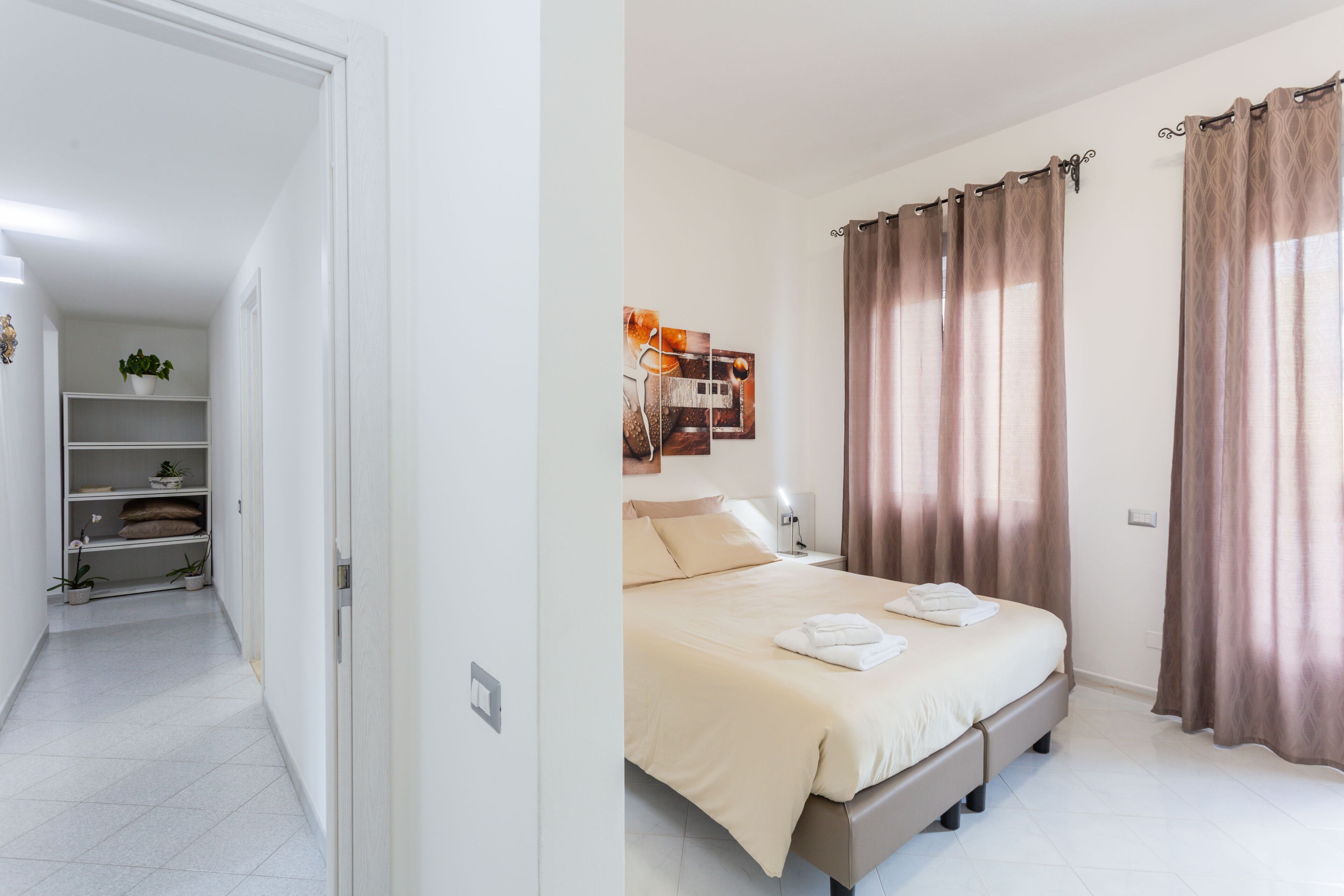 A hotel room with a bed, bedside table, and curtains. There is a hallway with a plant and a shelf.
