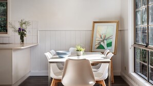 A dining area with a white table, white chairs, a vase with flowers, and a framed painting on the wall.
