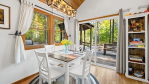 A dining area with a wooden table, white chairs, a vase with flowers, and a view of the forest through large windows. - A Relaxation Station (Morgantown)
