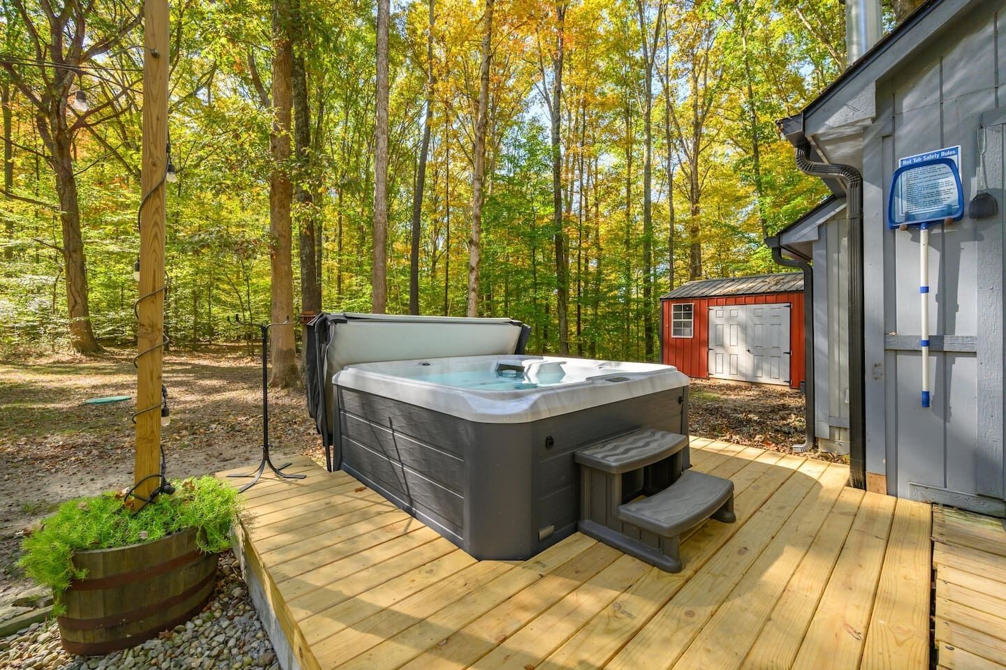 A hot tub on a wooden deck with a forest backdrop, a shed, and a safety sign.