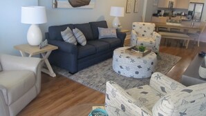 A living room with a dark blue sofa, a round ottoman with a starfish pattern, a white armchair, and a coffee table with a plant. There is a painting of a boat on the wall, and a kitchen area is visible in the background.