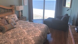 A bedroom with a large bed, a small sofa, a nightstand, a lamp, and a view of the sea through a large window.