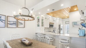A modern kitchen with white cabinets, a wooden ceiling, and a dining area with white chairs and a wooden table. - High Pointe Overlooking Lagoon Pool, Gulf Views! (Rosemary Beach)