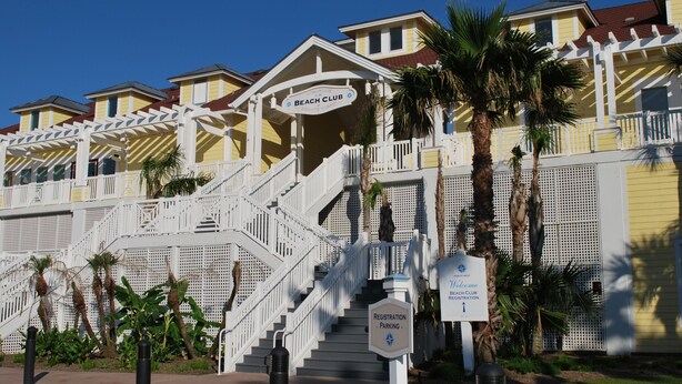 Un club de plage avec un escalier menant à l’entrée, des palmiers et un panneau indiquant le parking réservé aux membres.