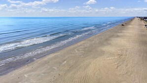 Une vaste plage de sable fin, avec des vagues douces, un ciel bleu clair et une côte lointaine.