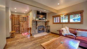 A cozy living room with a stone fireplace, wooden flooring, a leather sofa, and a large window with a view of the outside. - The Breckenridge Bicycle House - One Mile to Main St and the gondola (Breckenridge)