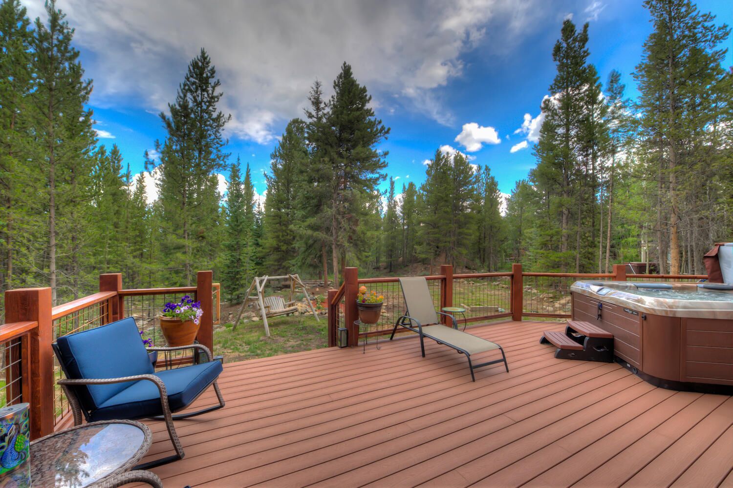 A wooden deck with a hot tub, lounge chairs, and a swing set overlooking a forest.