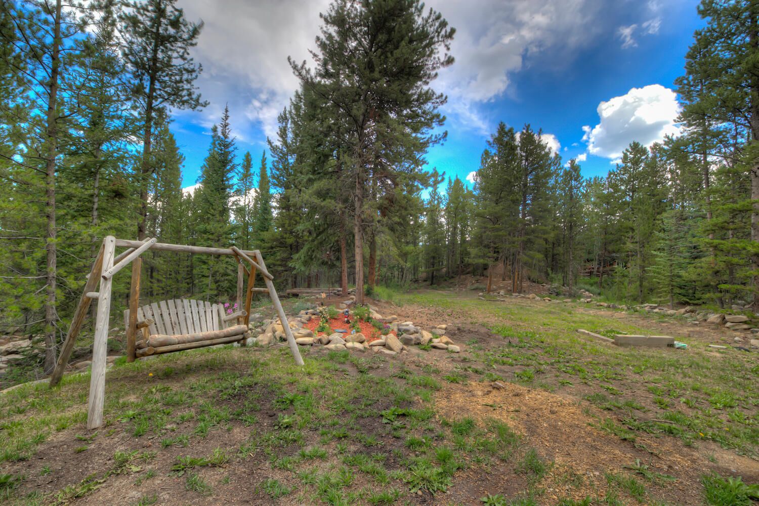 A wooden swing set in a forested area with a small rock garden and a clear sky.