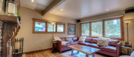 A spacious living room with a large sofa, a coffee table, and a fireplace. There are multiple windows providing natural light and views of the outdoors.