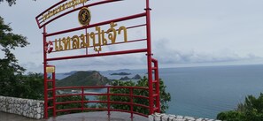 A red metal gate with Thai text and a logo, overlooking a coastal view with mountains and the sea.