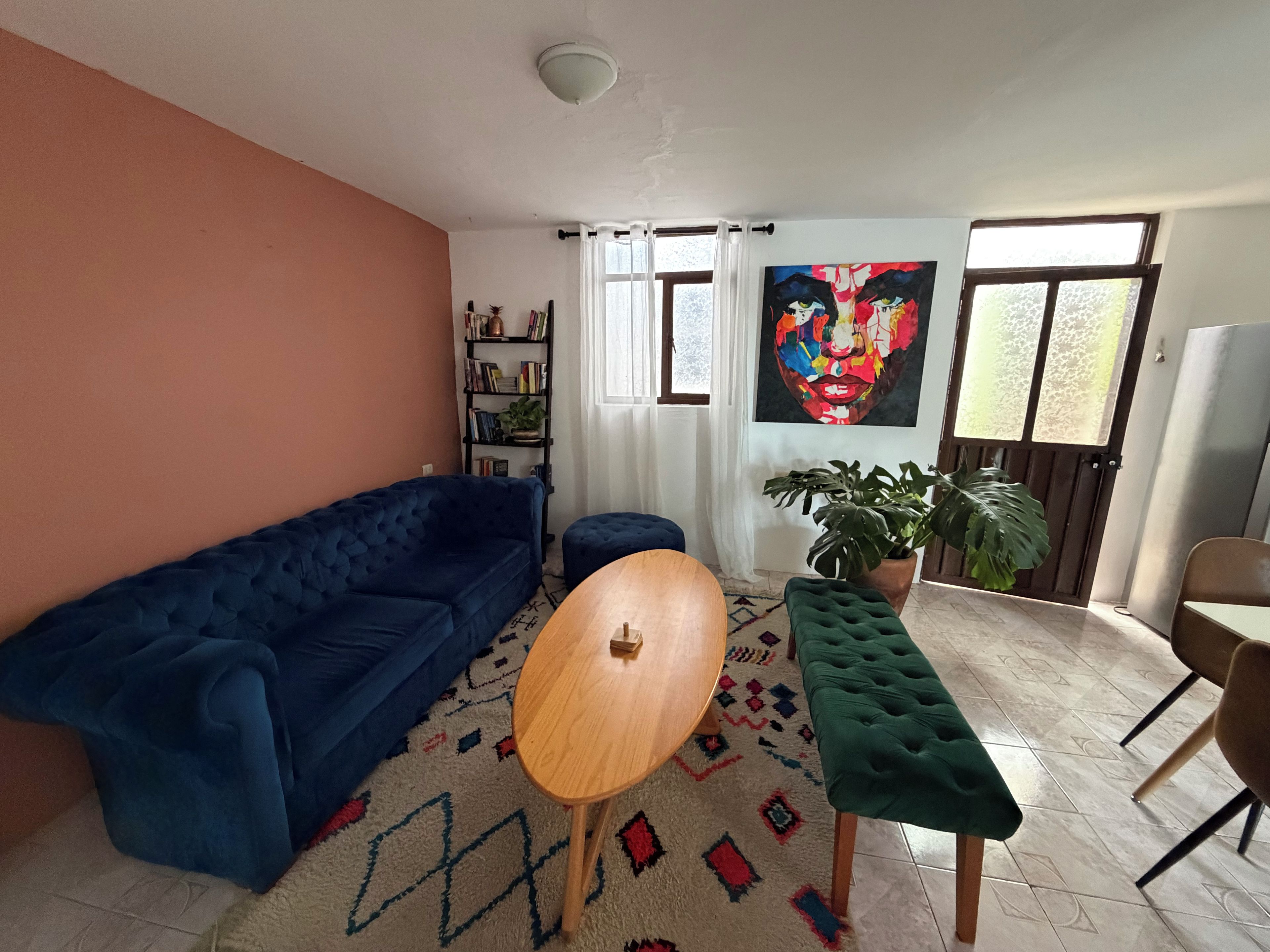 A living room with a blue sofa, a round wooden table, a green ottoman, a bookshelf, and a large abstract painting.
