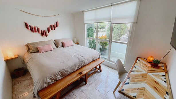 A bedroom with a large bed, a wooden bench, a chair, a small table, and a window with a view of greenery. - 2 Bed in the heart of Cholula with views to the Pyramid and the (San Andrés Cholula)