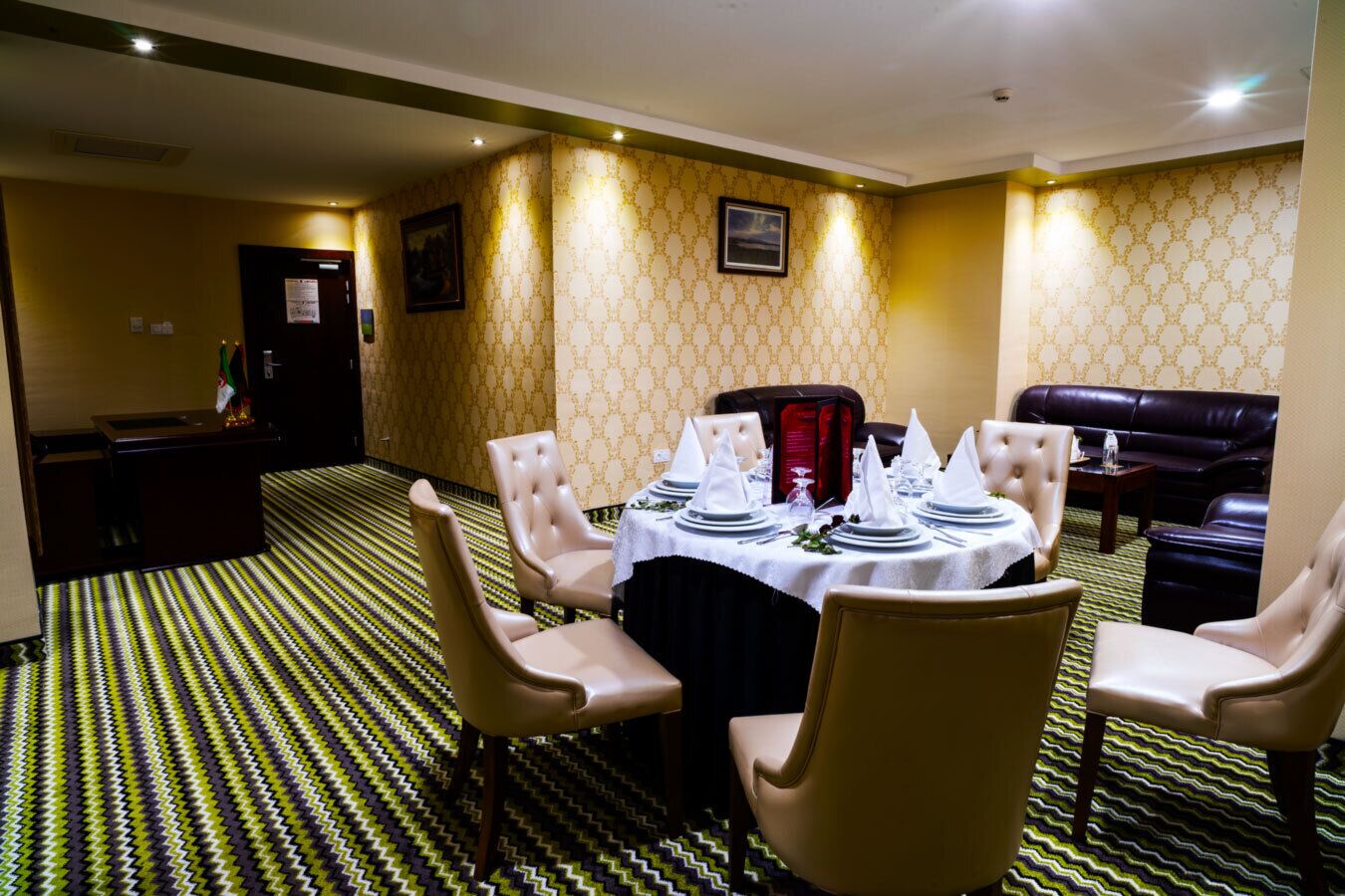 Un comedor con una mesa redonda preparada para cuatro personas, rodeada de cuatro sillas. Se observa un sofá de cuero negro y una pequeña mesa con una silla roja al fondo. La habitación tiene papel tapiz estampado y una alfombra con diseño en zigzag.