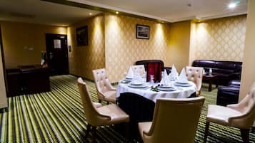 Un comedor con una mesa redonda preparada para cuatro personas, rodeada de cuatro sillas. Se observa un sofá de cuero negro y una pequeña mesa con una silla roja al fondo. La habitación tiene papel tapiz estampado y una alfombra con diseño en zigzag.