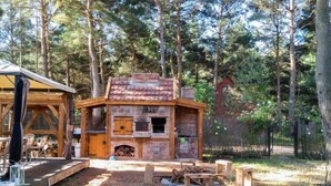 A rustic outdoor structure with a stone chimney, wooden benches, and a fire pit, surrounded by trees.