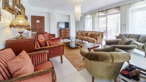 A spacious living room with a sofa set, a coffee table, and a television. There are decorative items on the coffee table and a chandelier hanging from the ceiling.