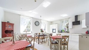 A spacious dining area with wooden chairs and tables covered in red and white checkered tablecloths, a kitchen area with white cabinets, and a large window providing natural light.