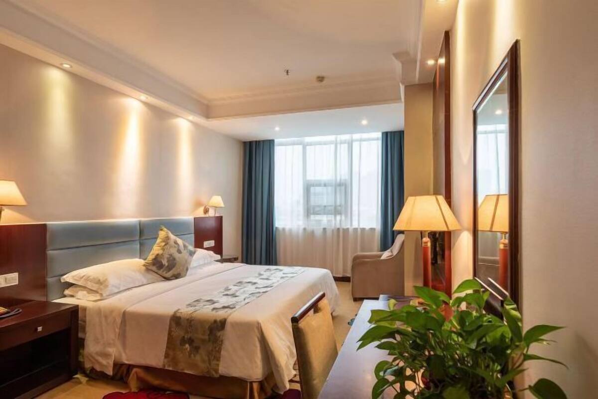 Una habitación de hotel con cama, mesitas de noche, una silla, un escritorio, un espejo y una planta.
