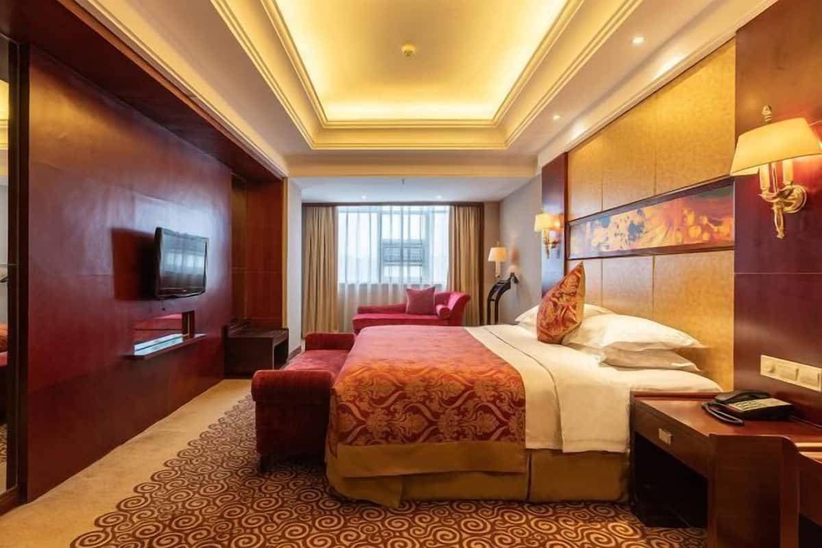 Habitación de hotel con una cama grande, un televisor montado en la pared, un escritorio con lámpara y una ventana con cortinas.