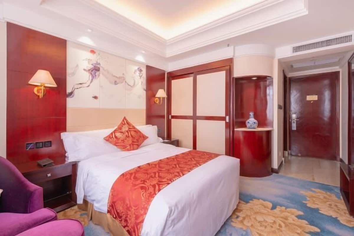 Una habitación de hotel con cama, mesita de noche, silla, un cuadro en la pared, una puerta y alfombra.