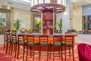Un hall de hotel con una barra circular central, varios ambientes de estar y plantas decorativas.
