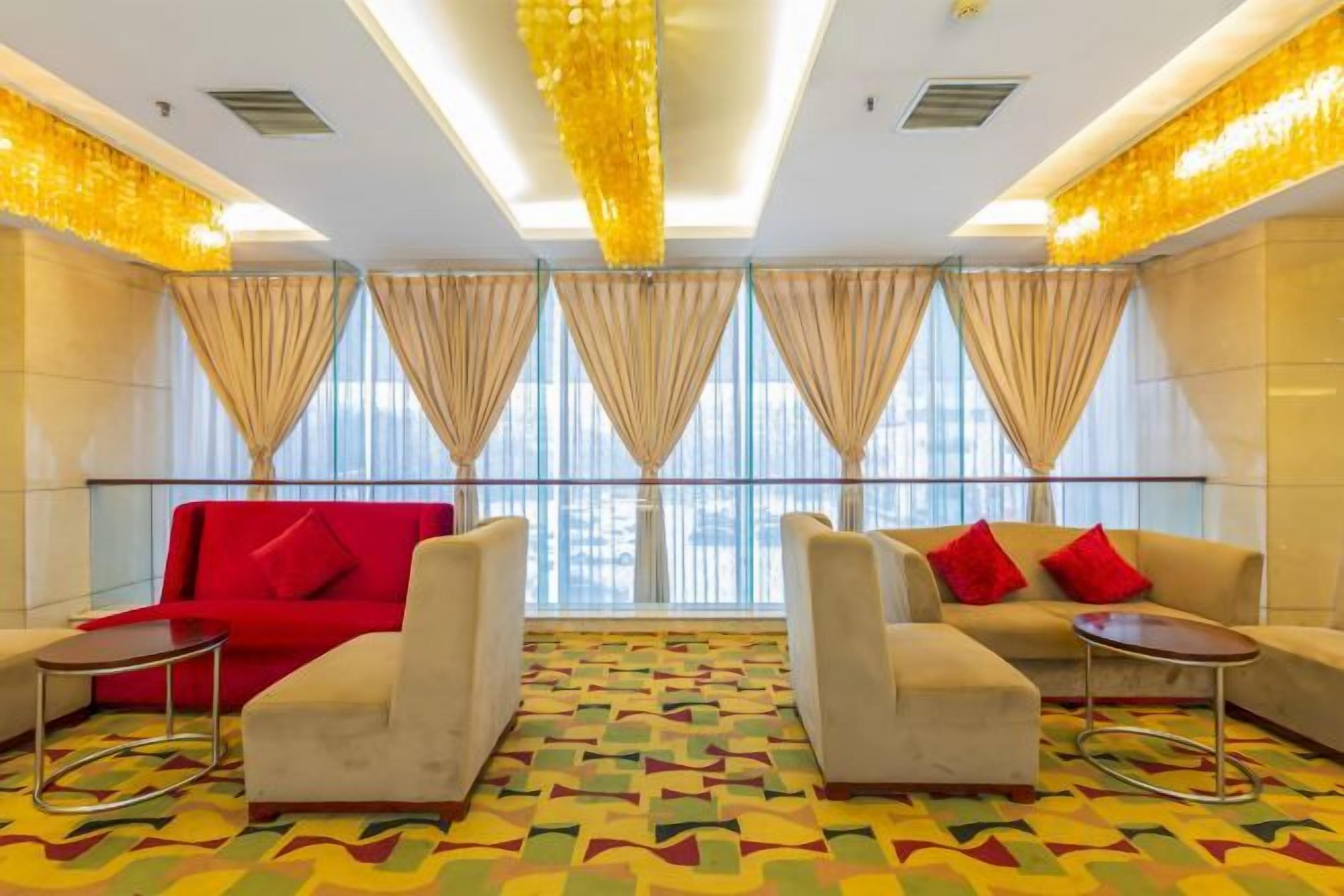 A hotel lobby with a red sofa, beige sofas, and armchairs, a patterned carpet, and large windows with curtains.